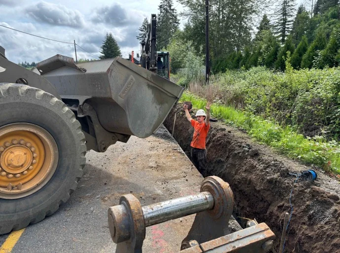 Worker standing in deep unprotected trench with excavator bucket overhead showing cave-in and struck-by hazards
