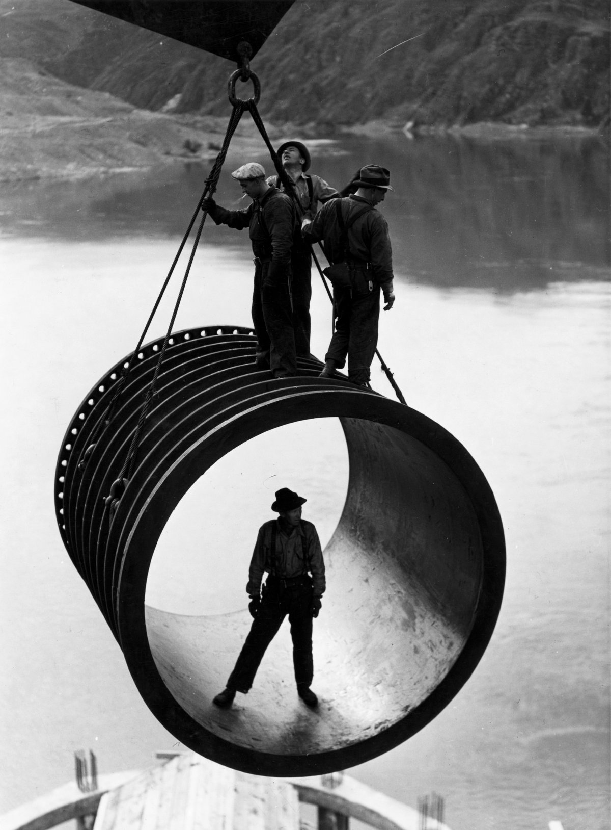Construction workers riding a crane-lifted pipe section during dam construction, 1930s